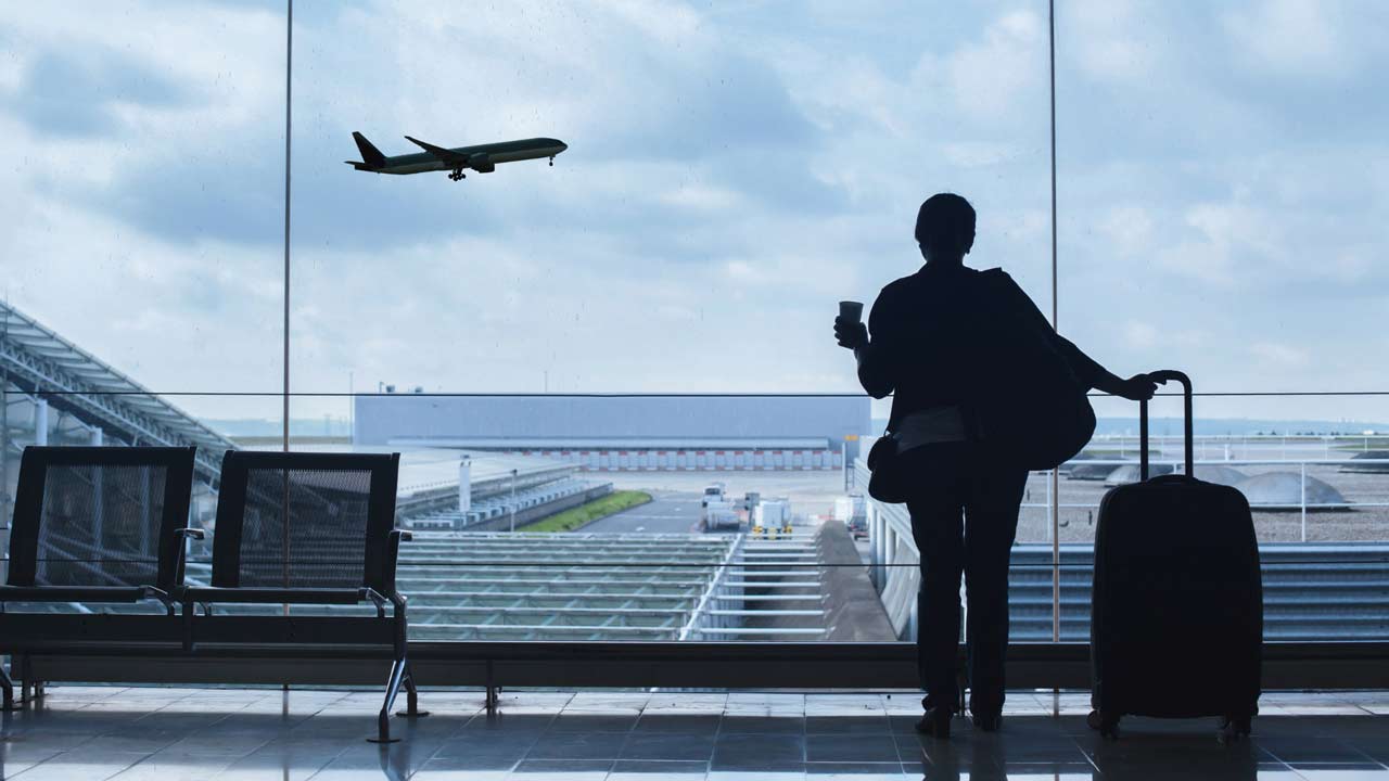Frau am Flughafen mit Koffer und Blick auf ein startendes Flugzeug